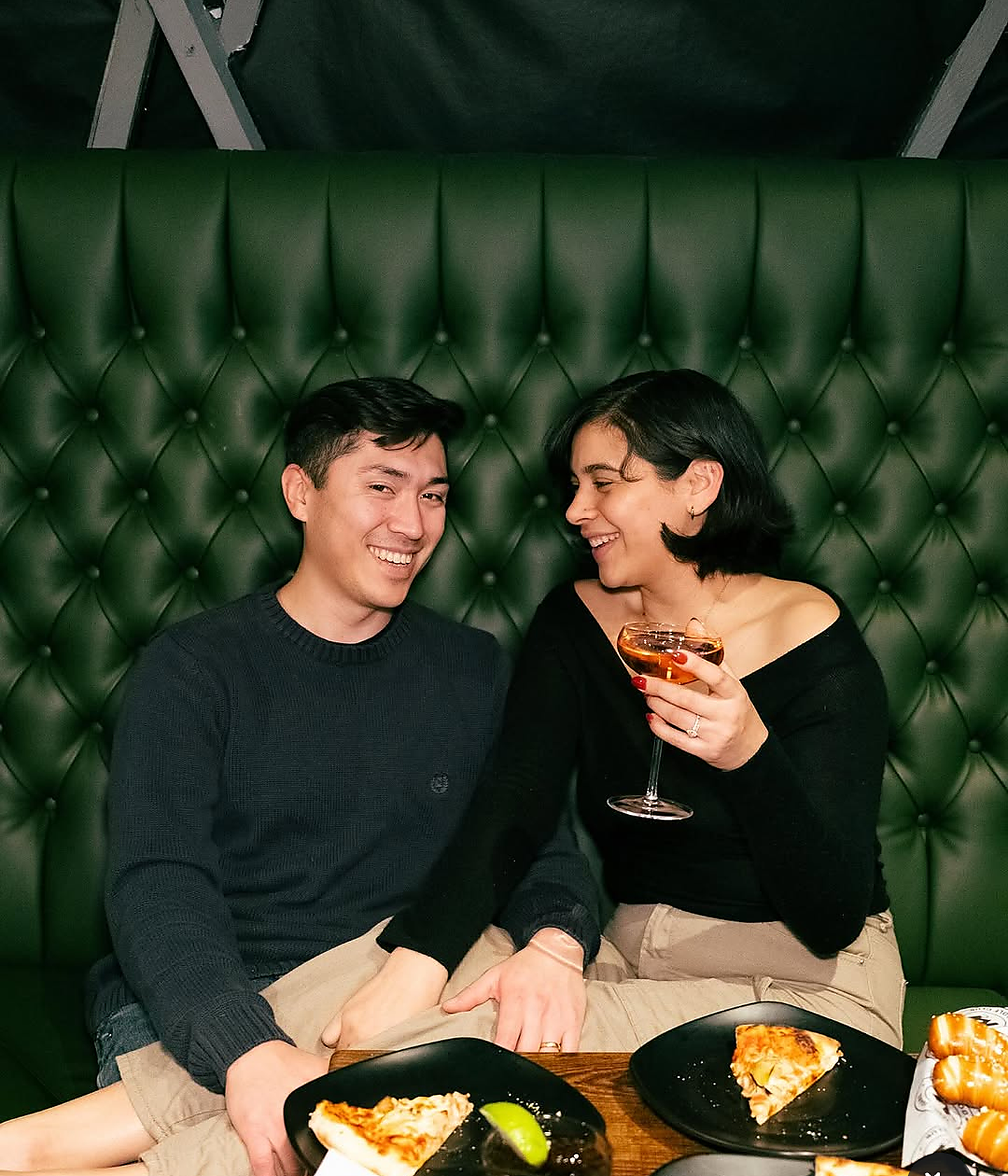 Couple laughing in deep green tufted leather booth enjoying craft cocktails in coupe glasses and pizza
